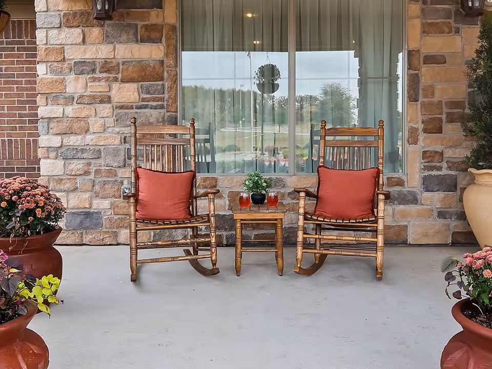 Two wooden rocking chairs with red cushions flank a small table holding plants and drinks on a stone-front porch with potted flowers.