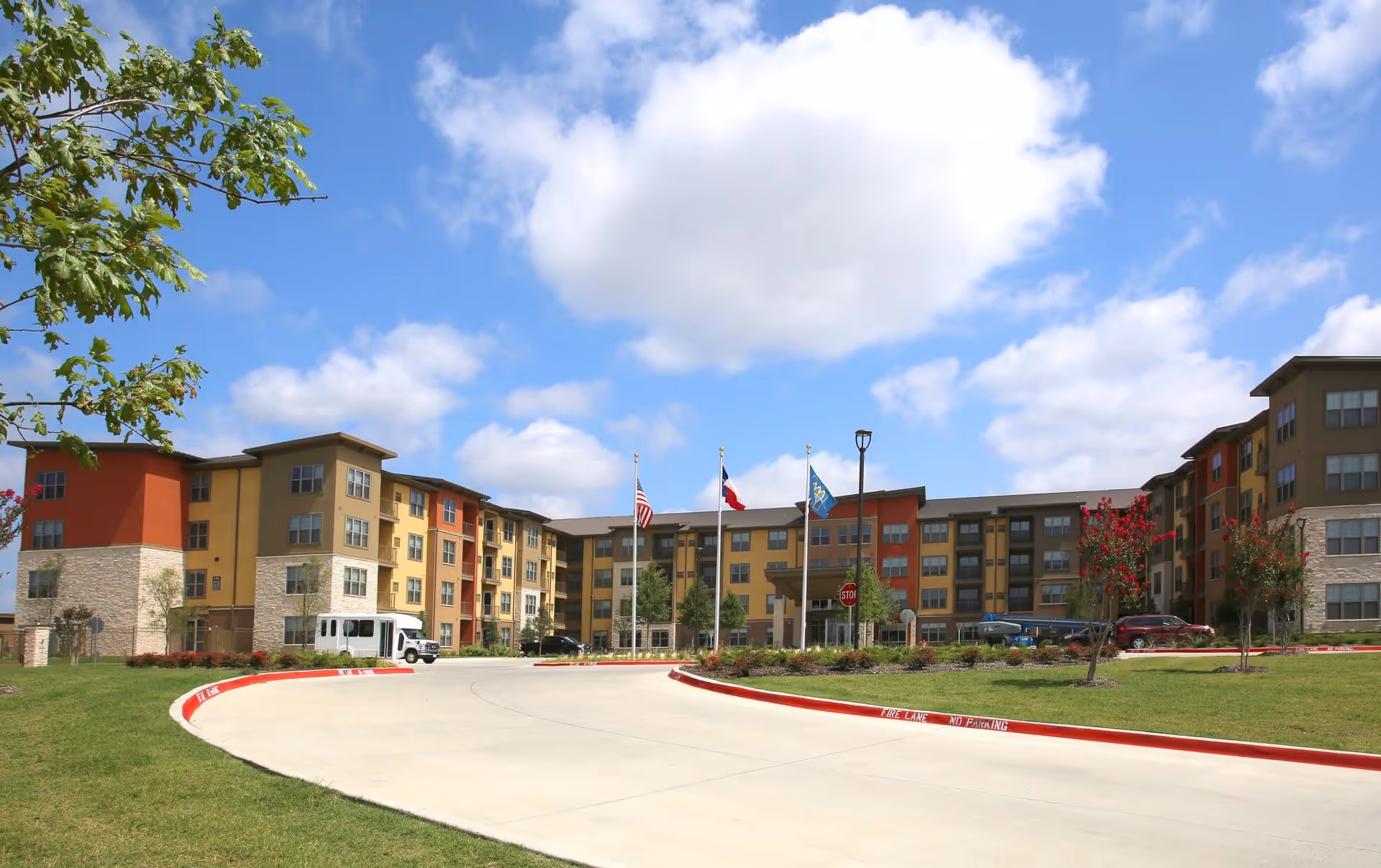 Front view of a multi-story senior living building with a circular driveway, flags, and landscaped grounds under a blue sky.