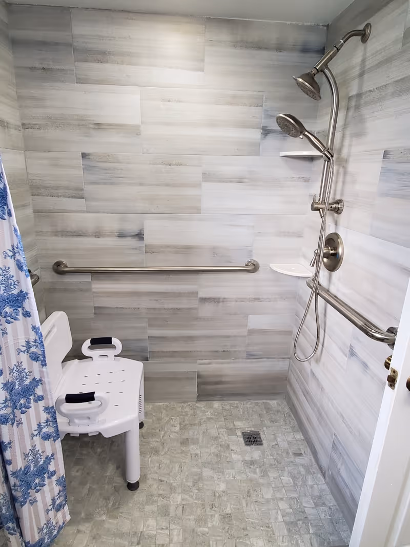 Accessible shower area with gray tiled walls and floor, equipped with a white plastic shower chair, two stainless steel grab bars, a dual shower head with handheld attachment, and a blue and white floral shower curtain partially visible on the left.