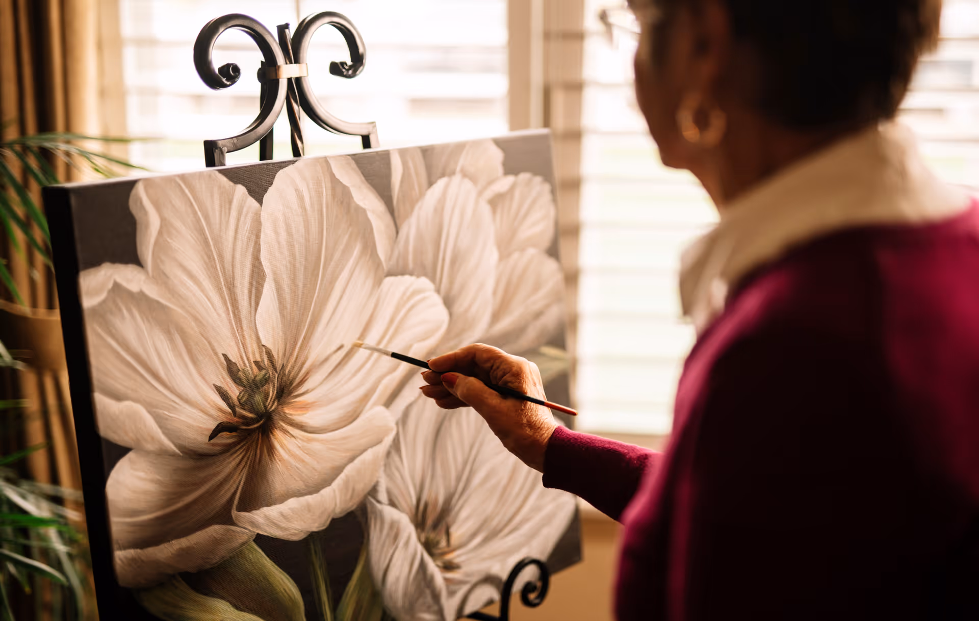 An elderly person painting a large white flower on a canvas placed on an easel in a warmly lit room with window blinds in the background.