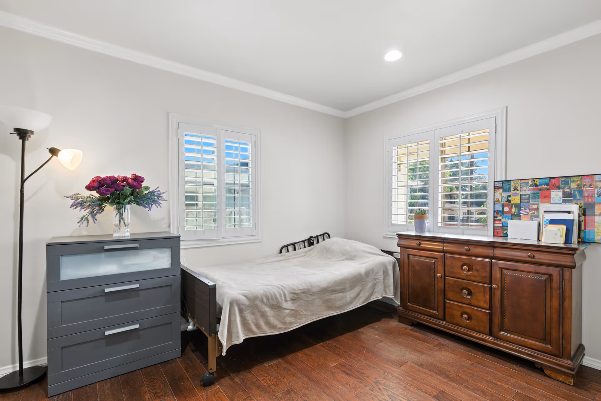 A compact resident bedroom with a bed, dresser, sideboard, and white-framed windows.