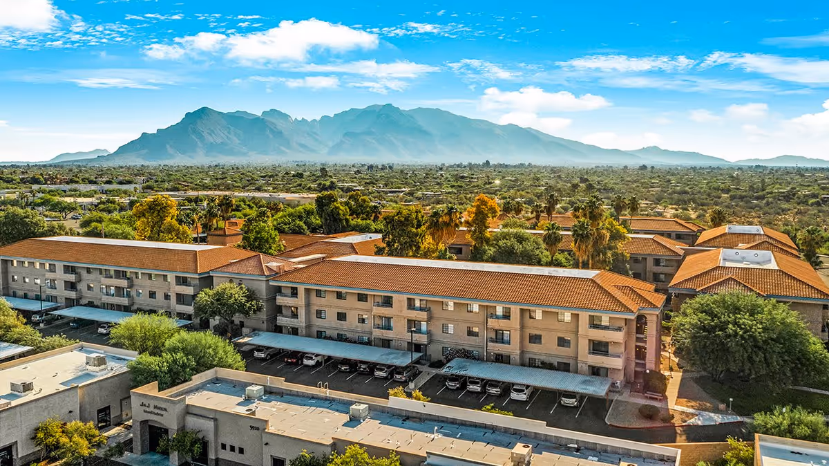 Aerial view of The Fountains at La Cholla senior living facility with multiple buildings featuring red-tiled roofs, surrounded by trees and desert landscape, with mountains in the background under a partly cloudy blue sky.