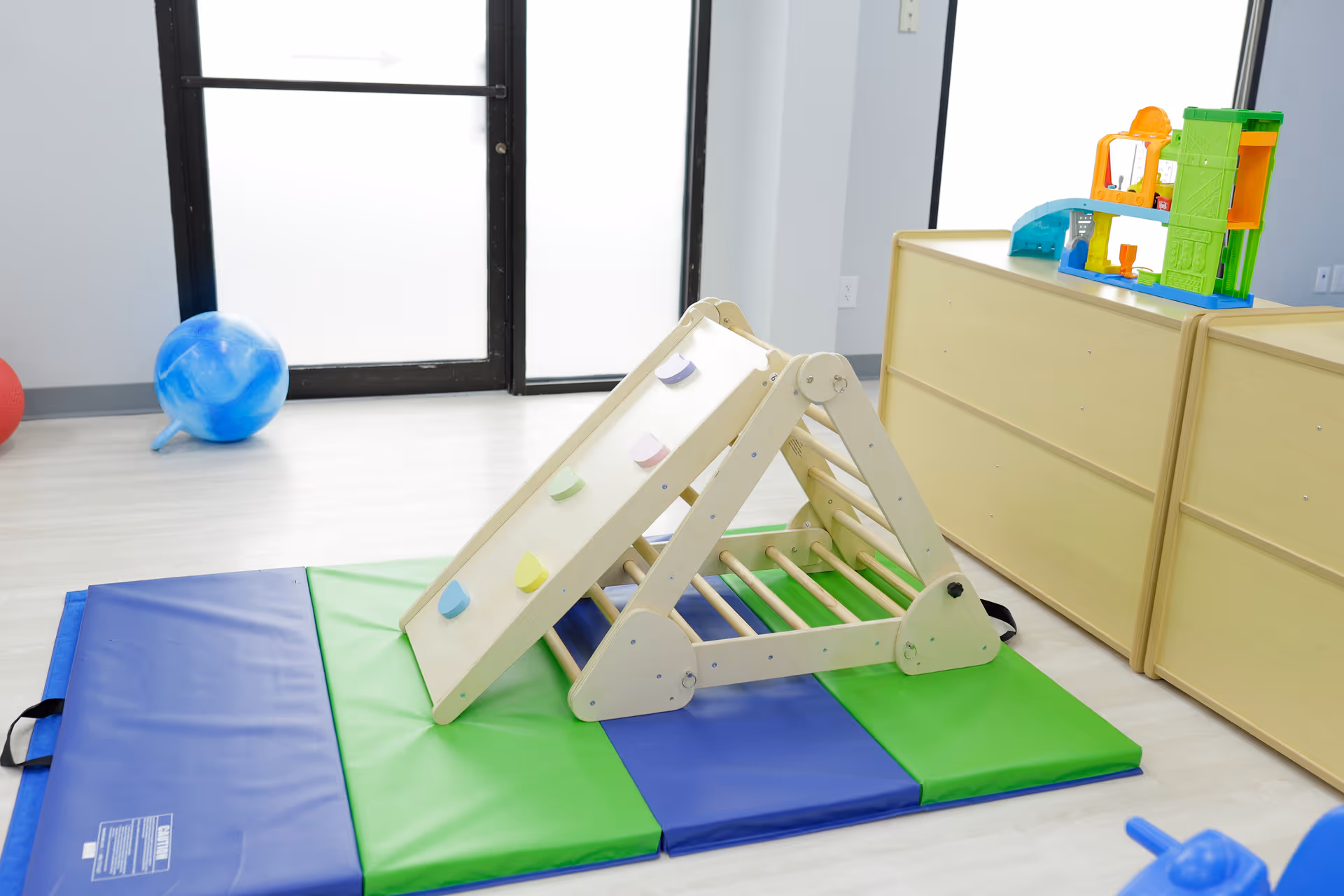 Indoor therapy playroom with a wooden climbing frame, mats, and colorful toys.