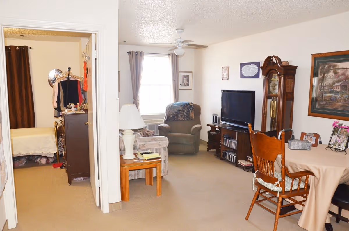 Interior view of a senior living facility showing a living room with a TV, armchair, sofa, wooden dining table with chairs, a grandfather clock, and framed pictures on the walls. To the left, an open doorway reveals a bedroom with a bed, dresser, and hanging clothes.