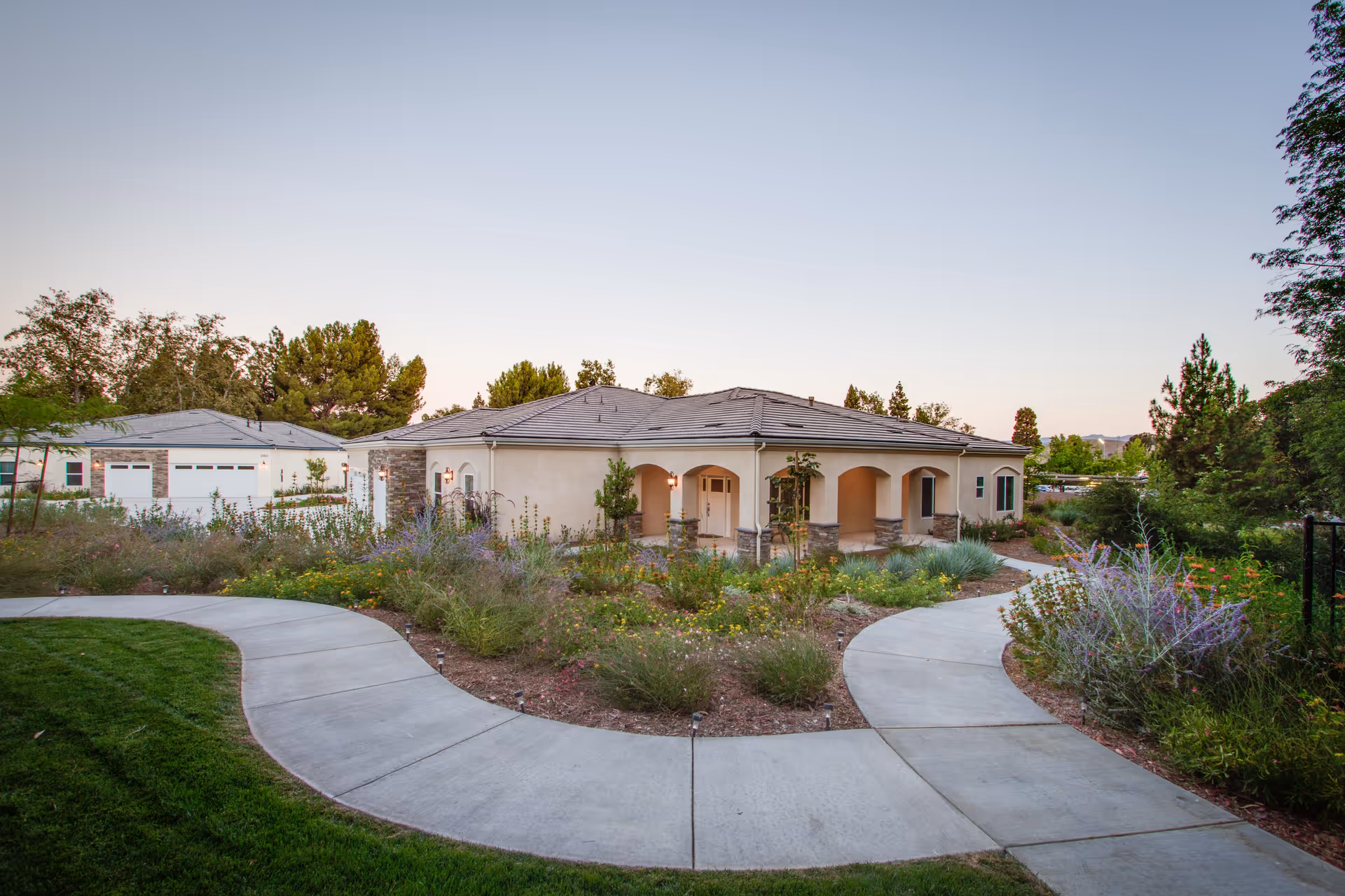 A single-story building with a tiled roof and arched porch surrounded by landscaped gardens and curved concrete walkways, set against a clear sky at dusk.
