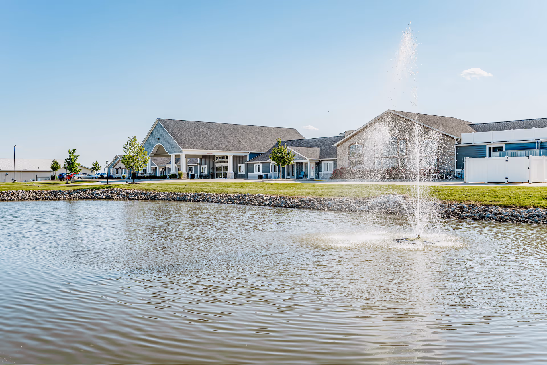 View of Villas of Holly Brook Assisted Living & Memory Care facility in Jerseyville, IL, showing a large pond with a water fountain in the foreground and the building with a pitched roof and multiple entrances in the background under a clear blue sky.