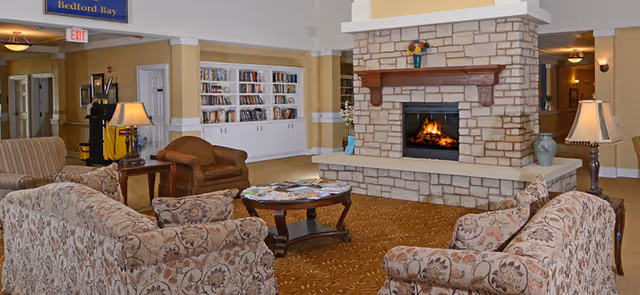 A cozy living room area in a senior living facility featuring a stone fireplace with a fire burning, surrounded by floral-patterned sofas and armchairs. A wooden coffee table with magazines sits in the center on a patterned carpet. Built-in white bookshelves filled with books are visible in the background, along with warm lighting and a sign that reads 'Bedford Bay'.