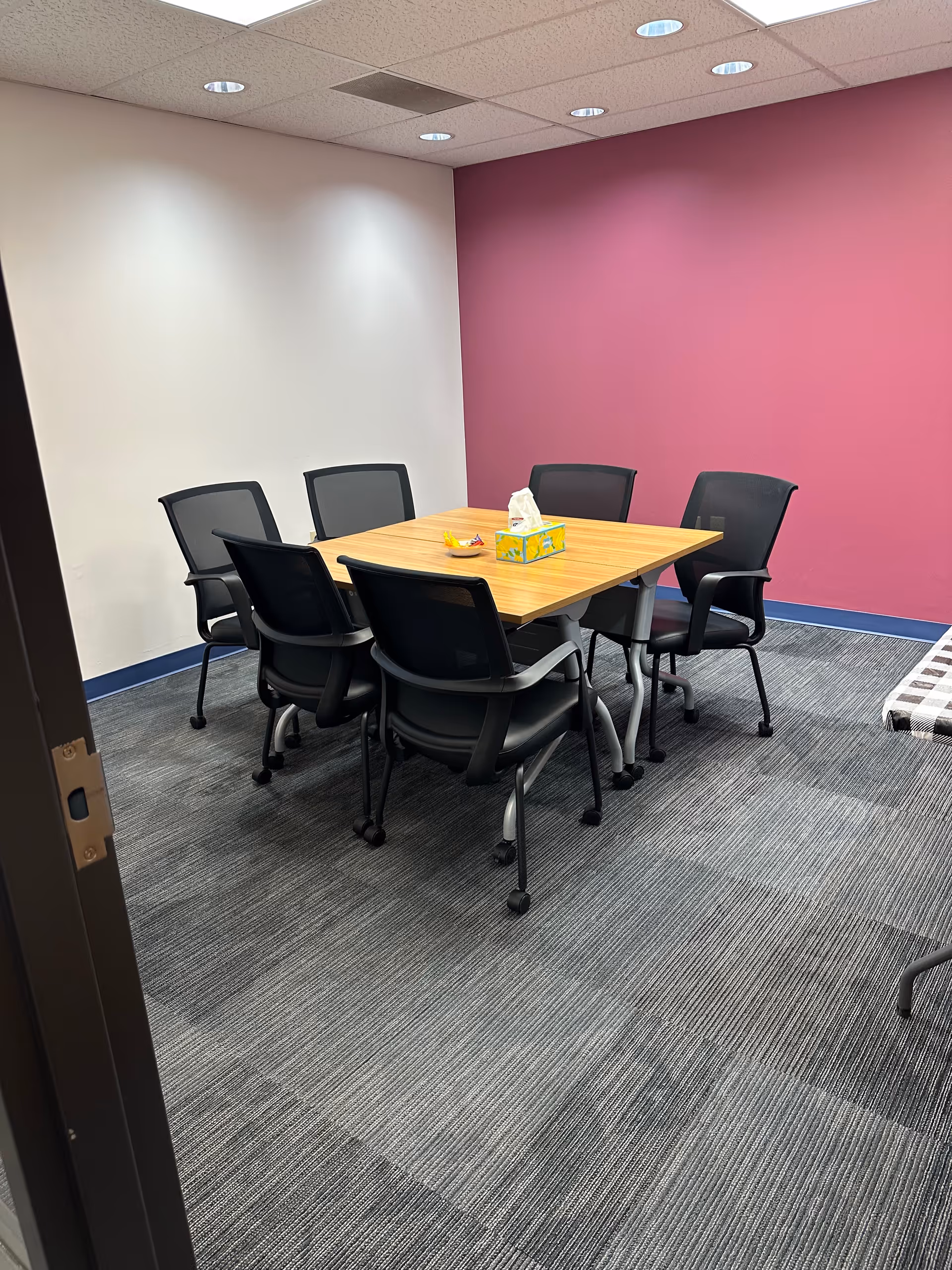A small dining and meeting room with a wooden table, black chairs, and magenta accent wall.