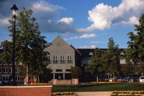 Exterior view of a senior living facility building with a triangular roof section, multiple windows, and surrounded by trees and landscaped flower beds under a partly cloudy sky.
