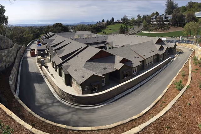 Aerial view of a single-story memory care building complex with a curved driveway and retaining walls on a hillside.