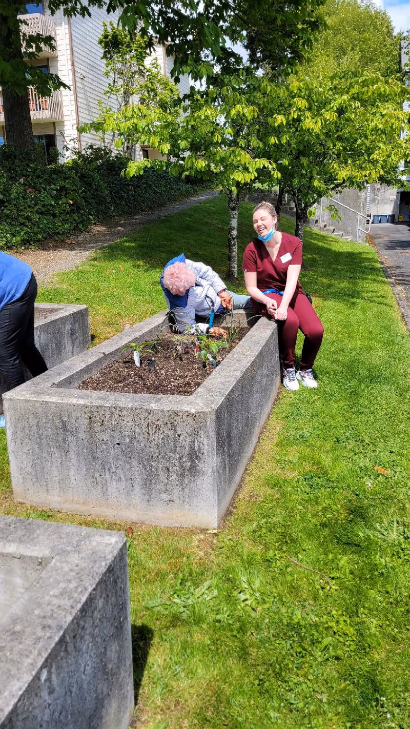 An elderly person and a caregiver are gardening together in a raised concrete planter outdoors on a sunny day. The caregiver, wearing a maroon uniform and a face mask pulled down, is sitting on the edge of the planter smiling. The elderly person is tending to the plants. There is green grass and trees around them, with a building partially visible in the background.