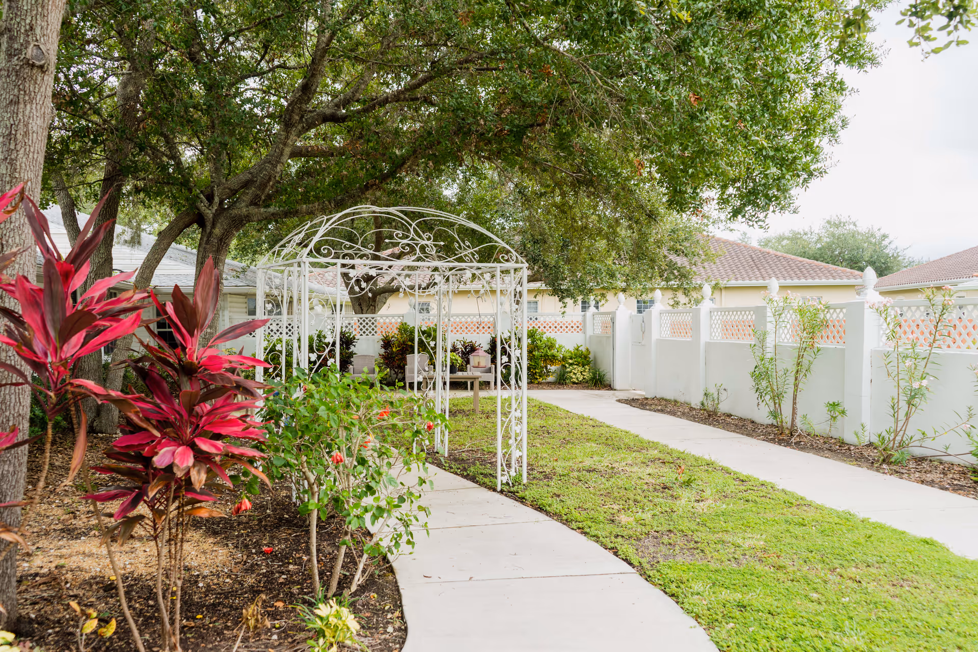 A garden pathway with a white metal archway surrounded by green trees and plants, including red-leafed shrubs. The path curves through a grassy area bordered by a white fence and leads to a seating area with chairs under the shade of large trees.