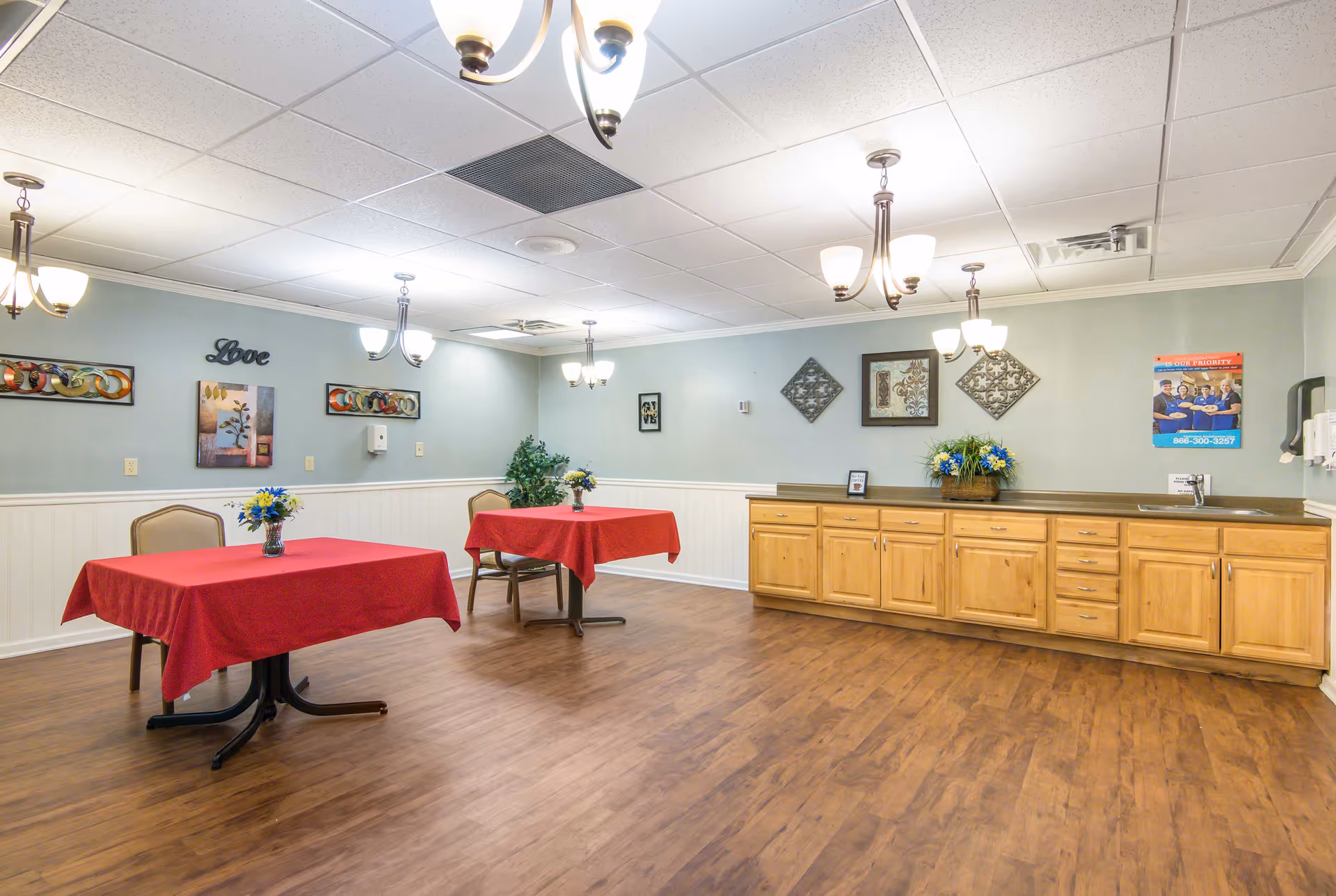 A spacious senior living dining area with red tablecloths, chairs, and wooden cabinets along the wall.