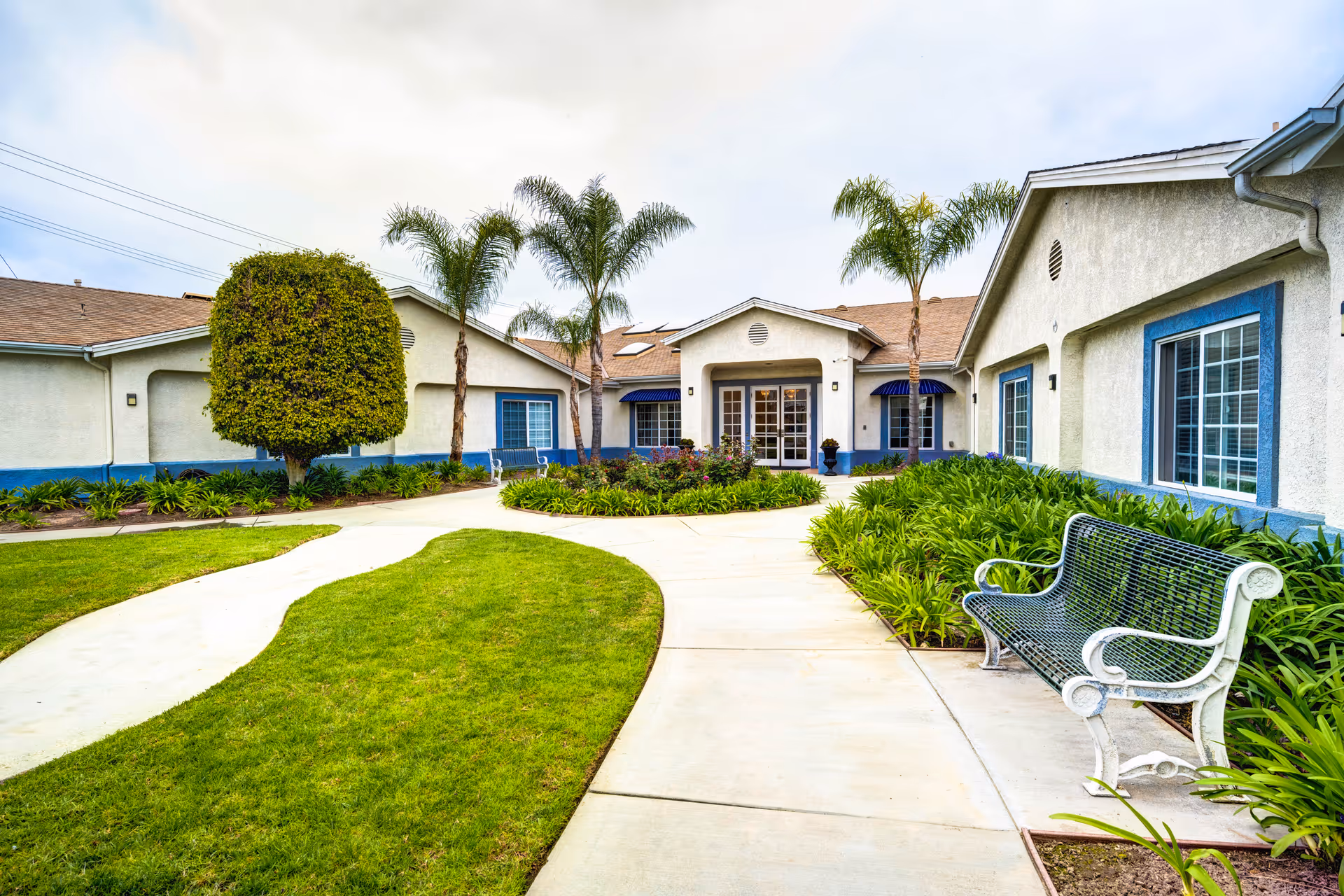 Exterior view of Ivy Terrace at Garden Grove Memory Care facility showing a pathway leading to the entrance. The building is single-story with white walls and blue trim around windows. There are palm trees, a neatly trimmed tree, green bushes, and a metal bench along the pathway.