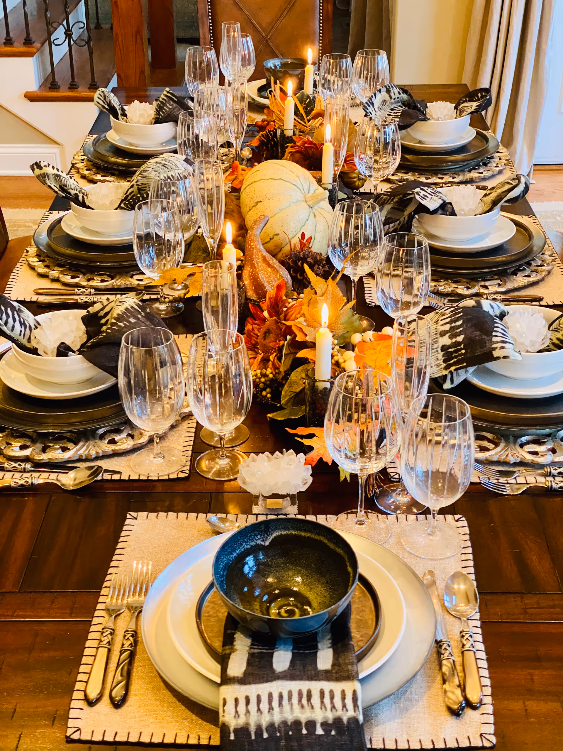 A dining table set for a meal with multiple place settings including plates, bowls, silverware, and glasses. The table is decorated with autumn-themed centerpieces featuring candles, pine cones, leaves, and a white pumpkin. Each place setting has a black and white patterned napkin and the table is in a warmly lit room with stairs visible in the background.
