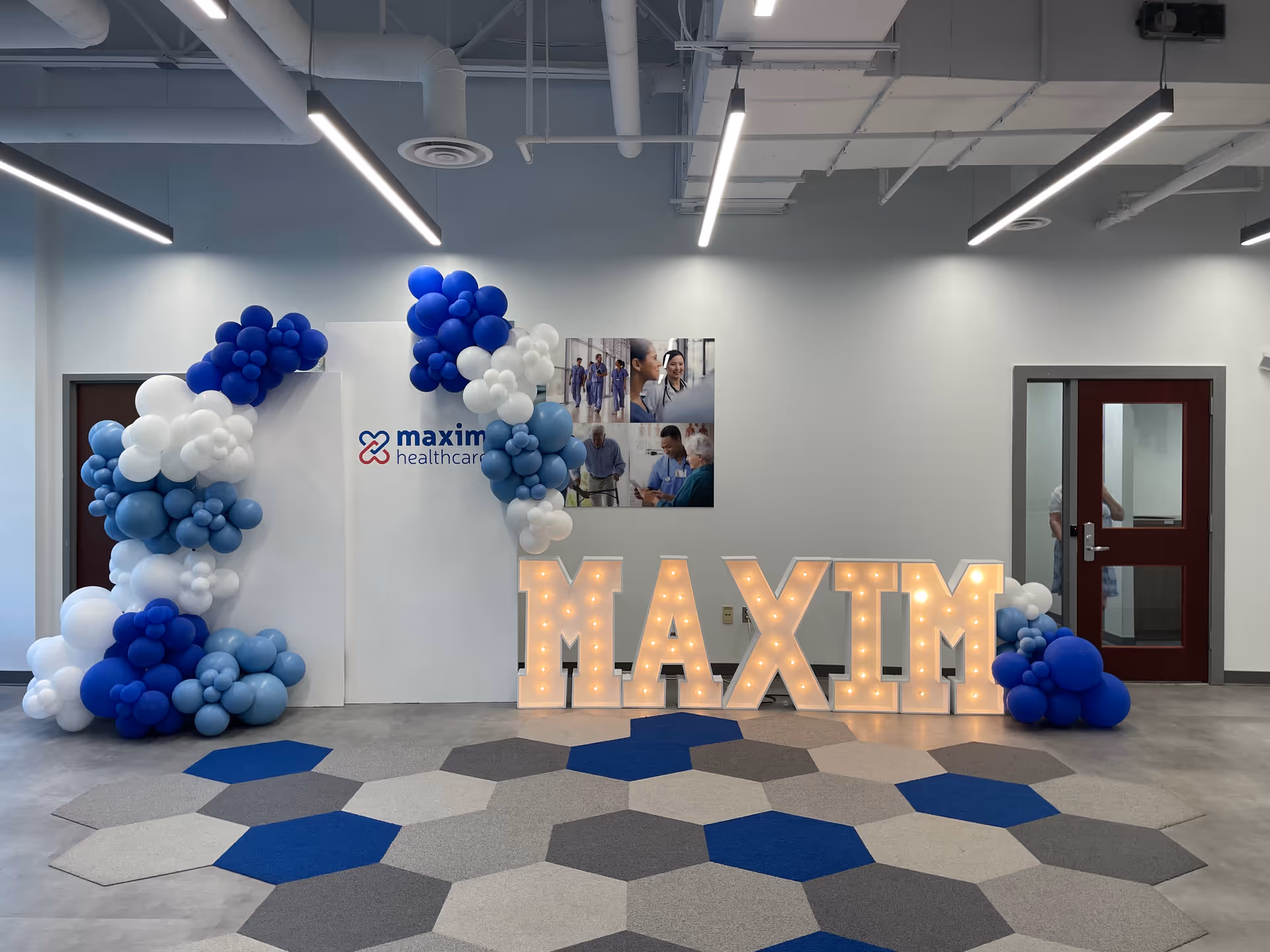 Indoor event space with illuminated MAXIM letters, blue and white balloons, and geometric floor tiles.