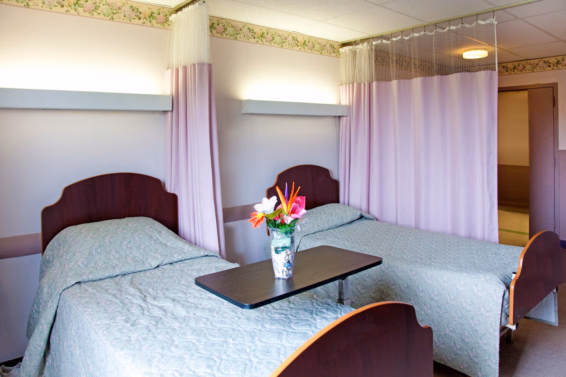 A room with two single beds separated by a privacy curtain. Each bed has a wooden headboard and is covered with a light green quilted bedspread. A small table with a vase of colorful flowers is placed between the beds. The walls are light-colored with a floral border near the ceiling, and there is a door visible in the background.