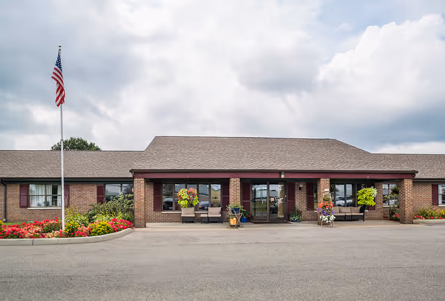 Front exterior view of a single-story brick building with a pitched roof, featuring a covered entrance with seating and potted plants. An American flag is displayed on a flagpole to the left, and flower beds with colorful flowers line the front of the building under a partly cloudy sky.