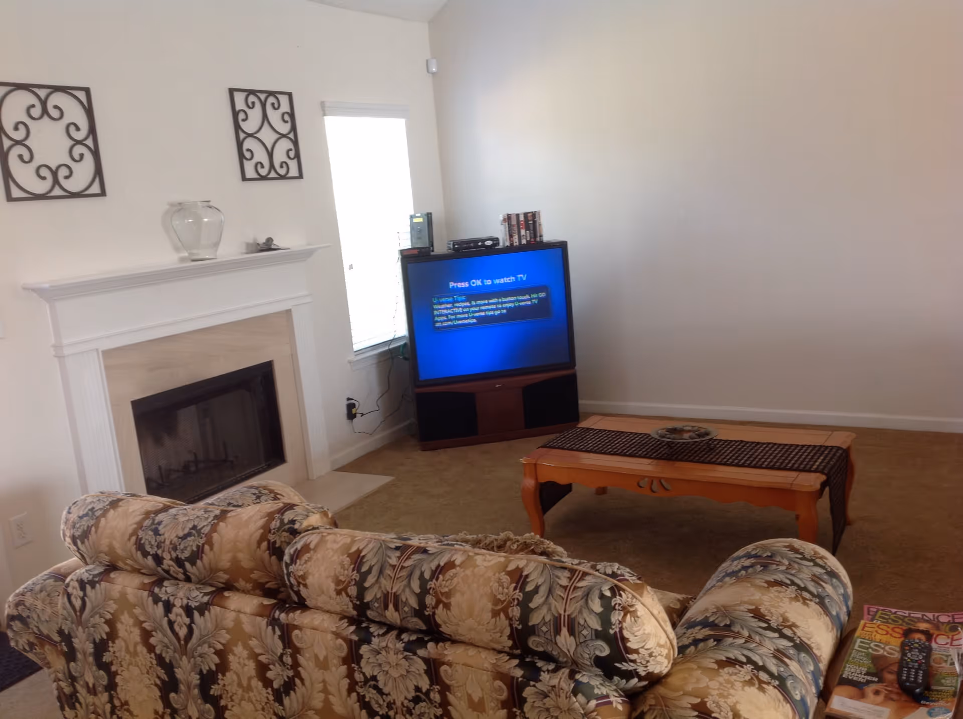 Living room with a floral-patterned sofa facing a fireplace, coffee table and a television against a blank wall.
