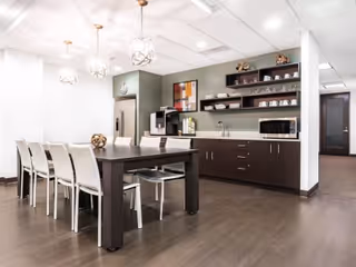 Modern senior living dining area with long dark table, white chairs, and kitchenette.
