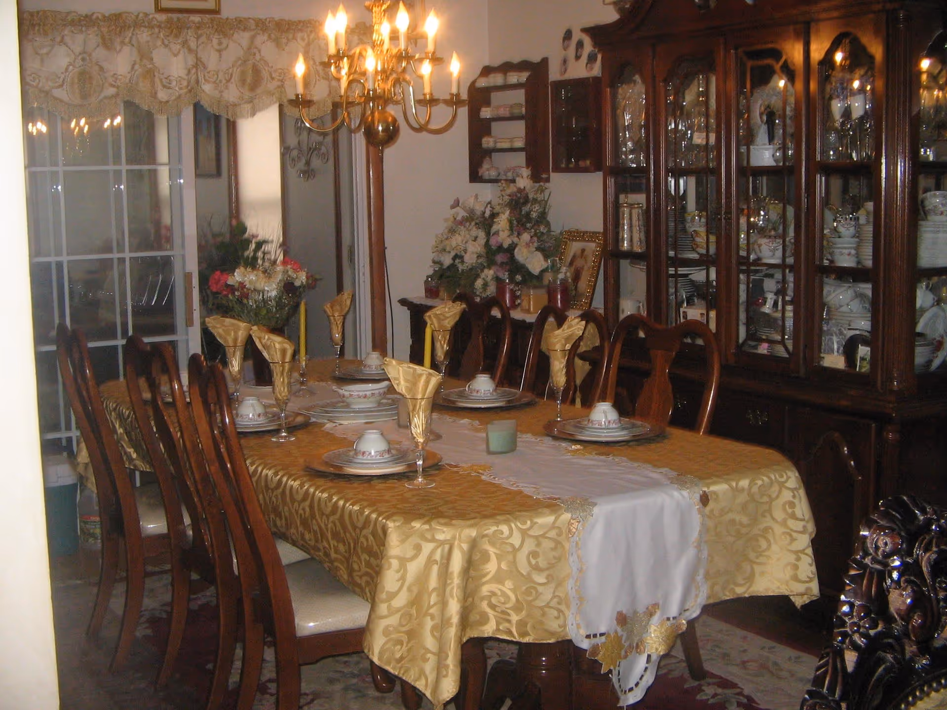 A formal dining room with a long wooden table covered with a gold patterned tablecloth and a white decorative runner. The table is set with plates, cups, and gold-colored napkins in glasses. There are eight wooden chairs around the table. A chandelier with lit candles hangs above the table. In the background, there is a wooden china cabinet filled with dishes and glassware, and floral arrangements on a sideboard. A sliding glass door with curtains is visible on the left side.