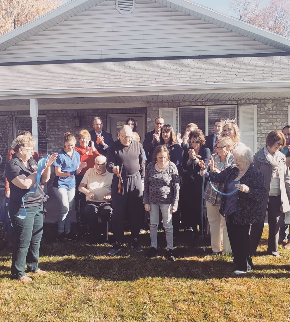 A group of people gathered on the lawn in front of a single-story house for a ribbon-cutting ceremony.