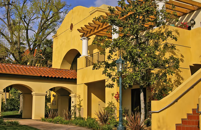 Exterior view of a yellow stucco building with arches, a red tile roof, a balcony with a pergola, and a staircase. There are trees, plants, and a vintage-style street lamp in front of the building under a clear blue sky.