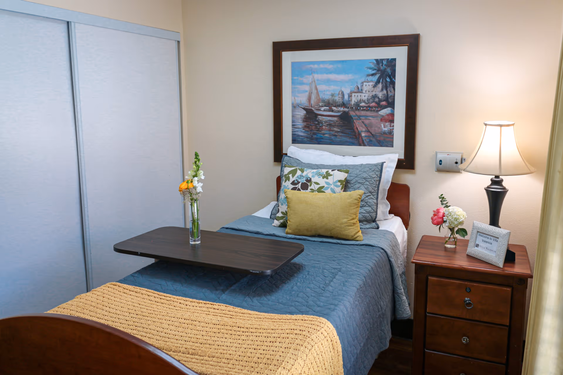 A neatly made single bed with blue bedding, multiple decorative pillows, and a yellow throw blanket at the foot. A dark wooden overbed table holds a small vase with flowers. Next to the bed is a wooden nightstand with a lamp, a small vase with flowers, and a framed sign. A large framed painting of a waterfront scene hangs on the wall above the bed. Sliding closet doors are visible on the left side of the room.