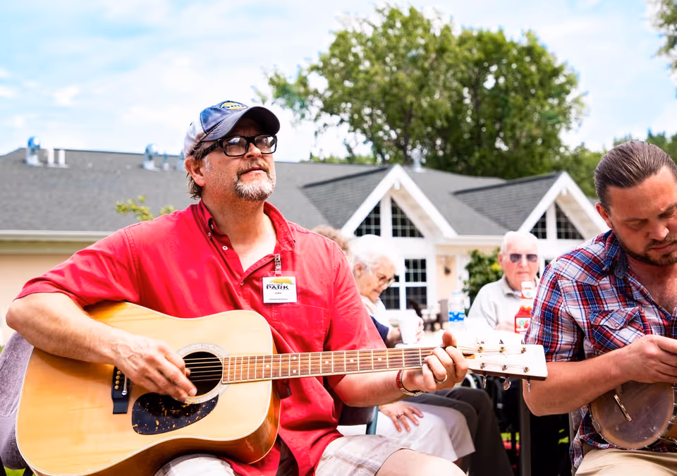 A man wearing a red shirt and a cap is playing an acoustic guitar outdoors. Another man in a plaid shirt is playing a banjo beside him. In the background, elderly people are seated and appear to be enjoying the music in a garden area with a building and trees behind them.
