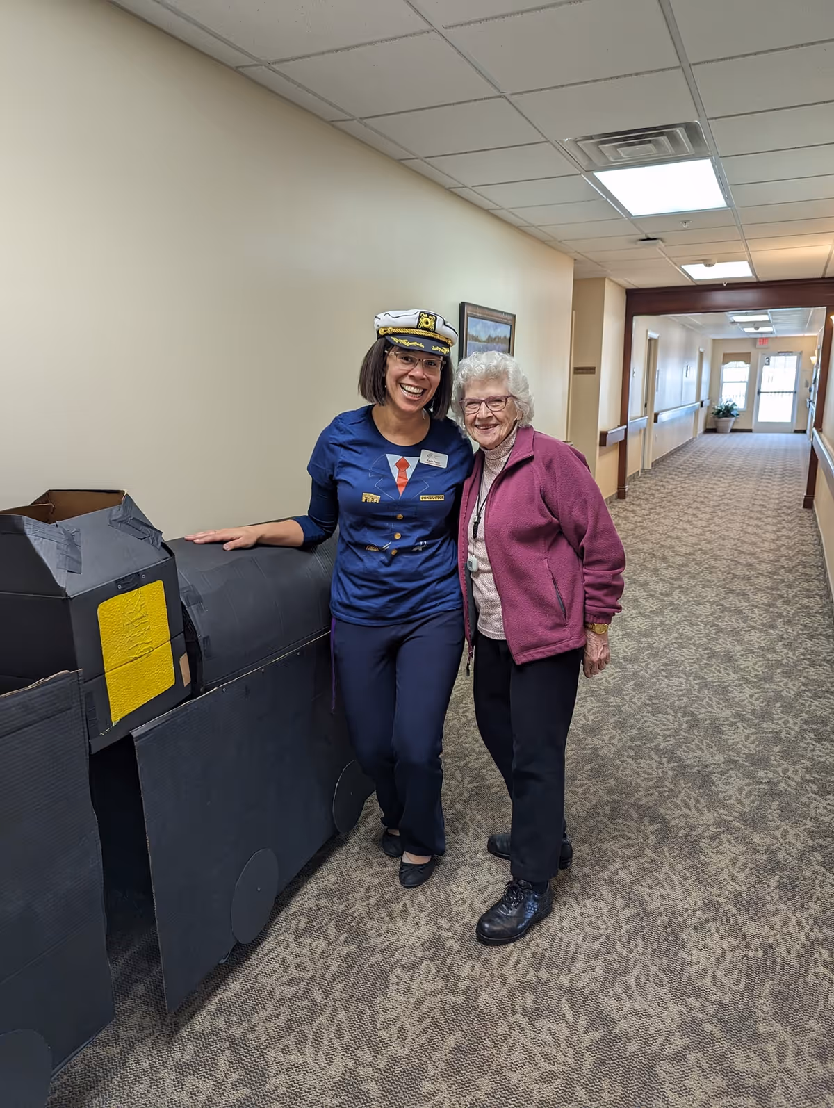 Two women standing in a carpeted hallway of an assisted living facility. One woman is dressed in a navy blue outfit with a captain's hat and is smiling, while the other is an elderly woman wearing glasses, a pink jacket, and black pants, also smiling. They are standing next to a large black cardboard structure resembling a train engine.