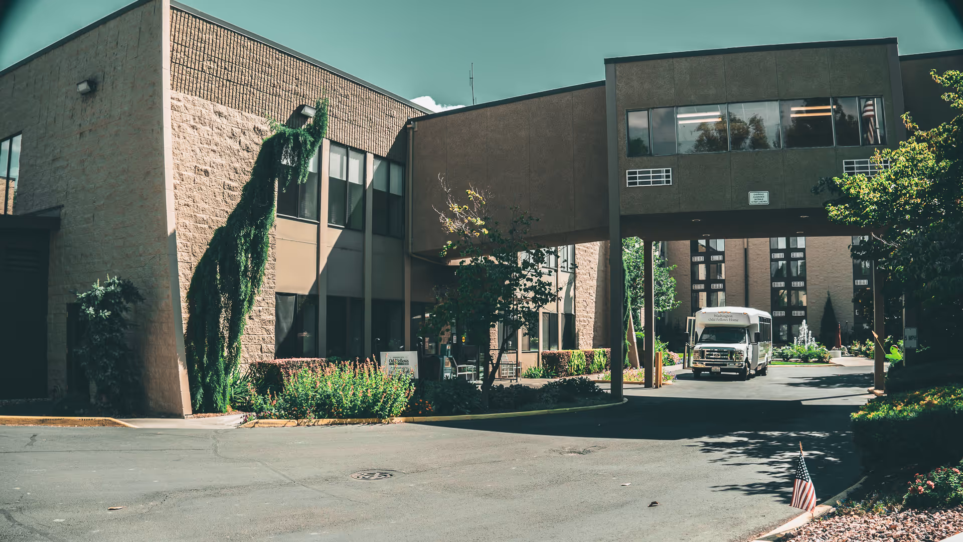 Exterior view of the Washington Odd Fellows Home building with a covered driveway and a shuttle bus parked underneath. The building is surrounded by greenery and trees, with a small American flag planted in the ground near the driveway.