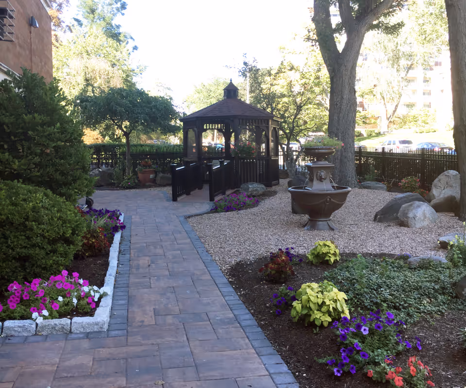 Paved walkway leading to a gazebo in a landscaped courtyard with a fountain and flower beds.