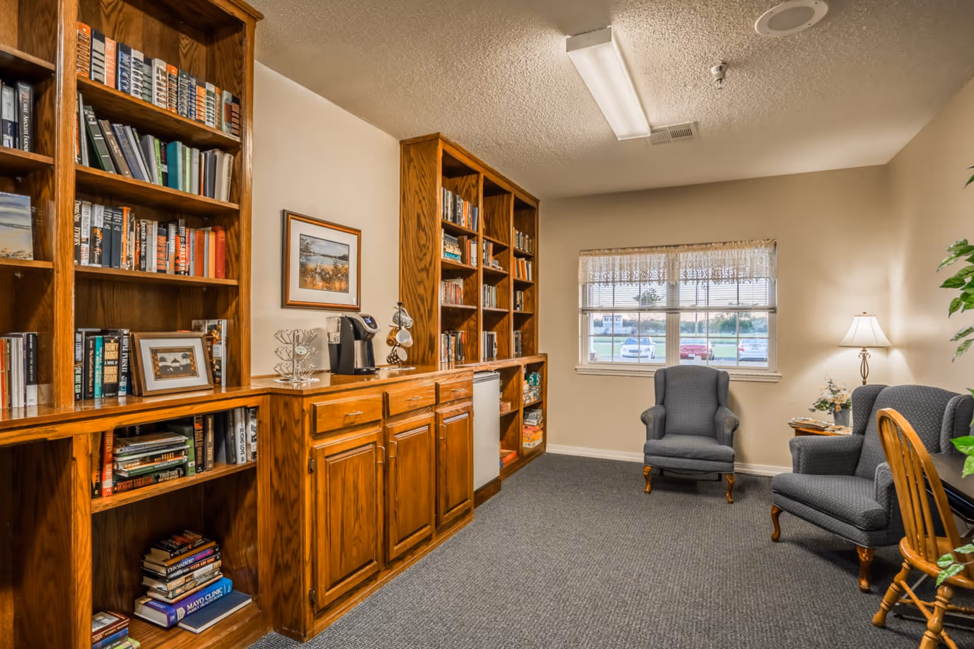 A cozy room with wooden bookshelves filled with books and a coffee maker on the counter. Two upholstered armchairs are positioned near a window with lace curtains, and a wooden chair is partially visible. The room has beige walls, a carpeted floor, and a table lamp providing warm light.