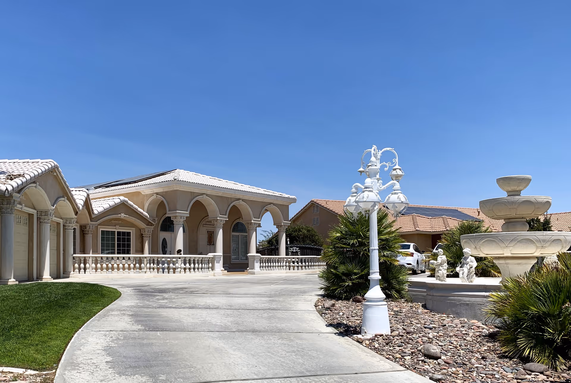 Front exterior of a Mediterranean-style single-story building with arched columns, a curved driveway, decorative lamp post and fountain under a clear blue sky.