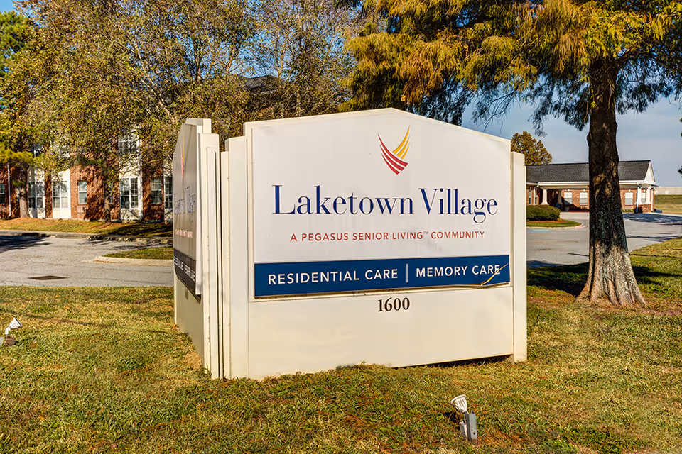 Large white sign reading 'Laketown Village' in front of the senior living campus with buildings and trees behind it.