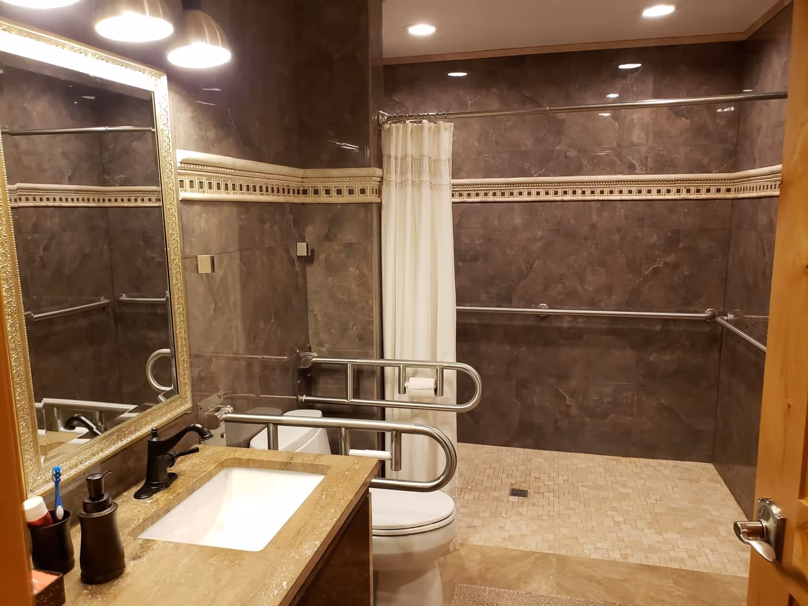 A bathroom with brown tiled walls and beige tiled floor. It features a sink with a countertop, a large mirror with a decorative frame, a toilet with safety grab bars, and a walk-in shower area with a white shower curtain and additional grab bars.