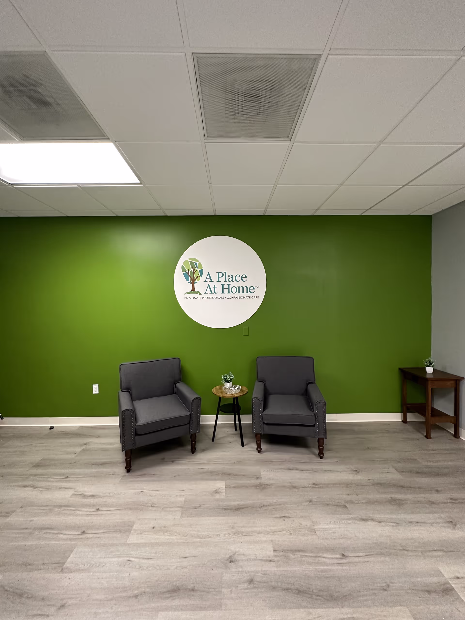 Two gray armchairs with a small table sit before a green wall and circular logo.