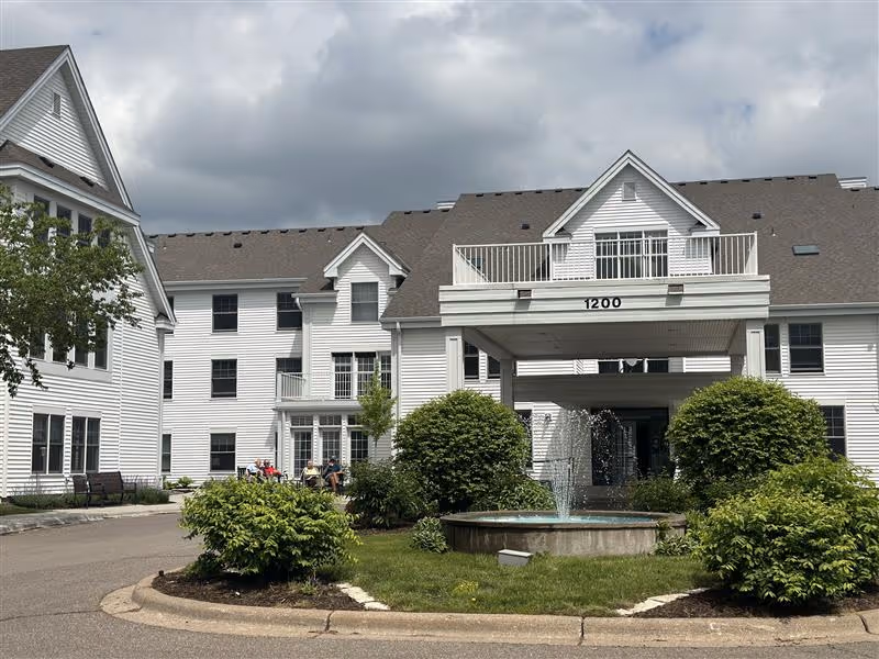 Ecumen Lakeview Commons exterior with white siding, circular driveway, fountain, and landscaped shrubs.
