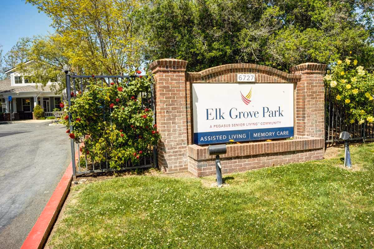 Brick entrance sign and gated entry for Elk Grove Park assisted living and memory care amid trees and a lawn.
