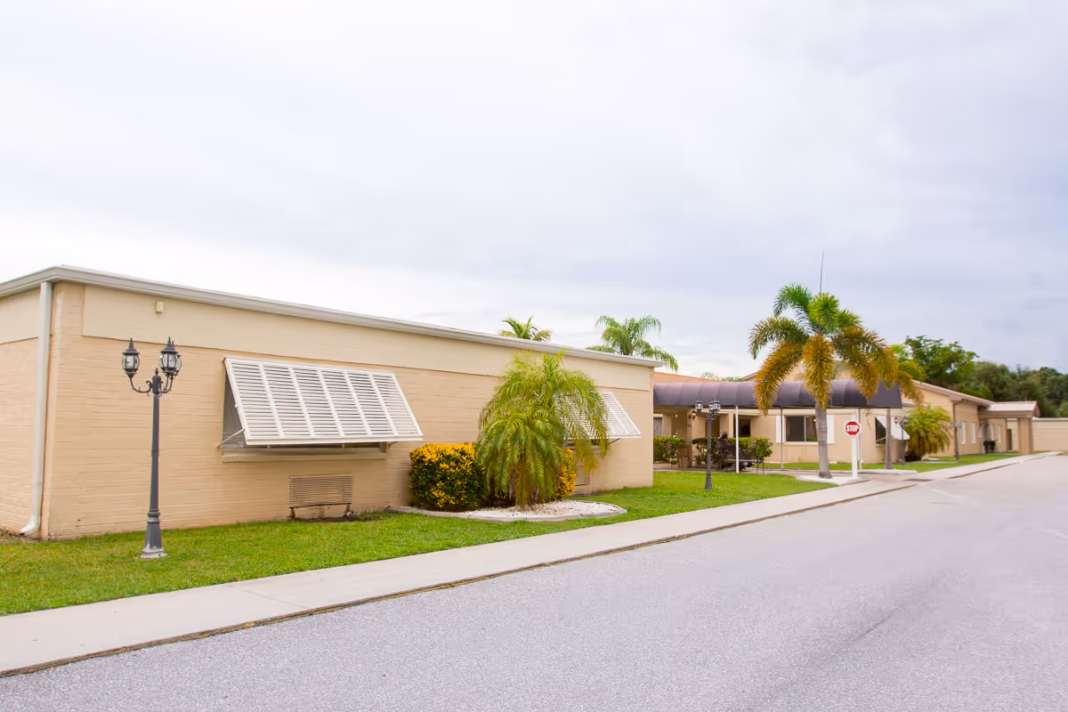 Exterior view of a single-story beige building with awning-covered windows, palm trees, a street lamp, and a stop sign along a paved road under a cloudy sky.