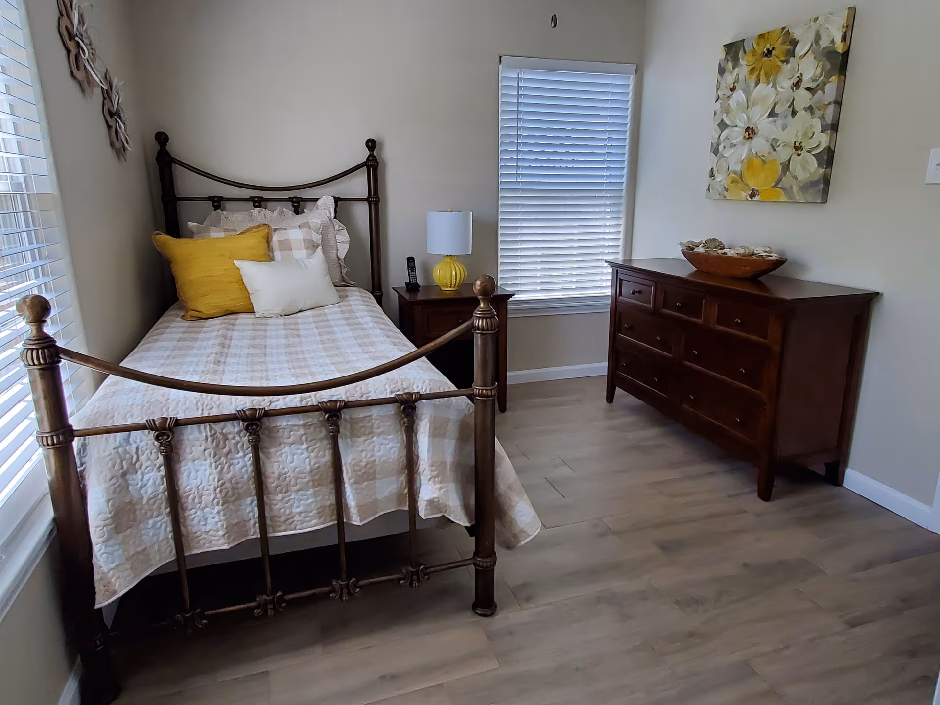 Sunlit senior living bedroom with a metal bed, nightstand, dresser, and wall art.