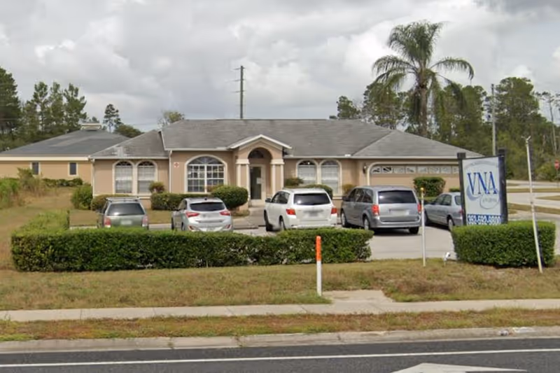 Single-story beige nursing facility with parked cars and a VNA sign in front.