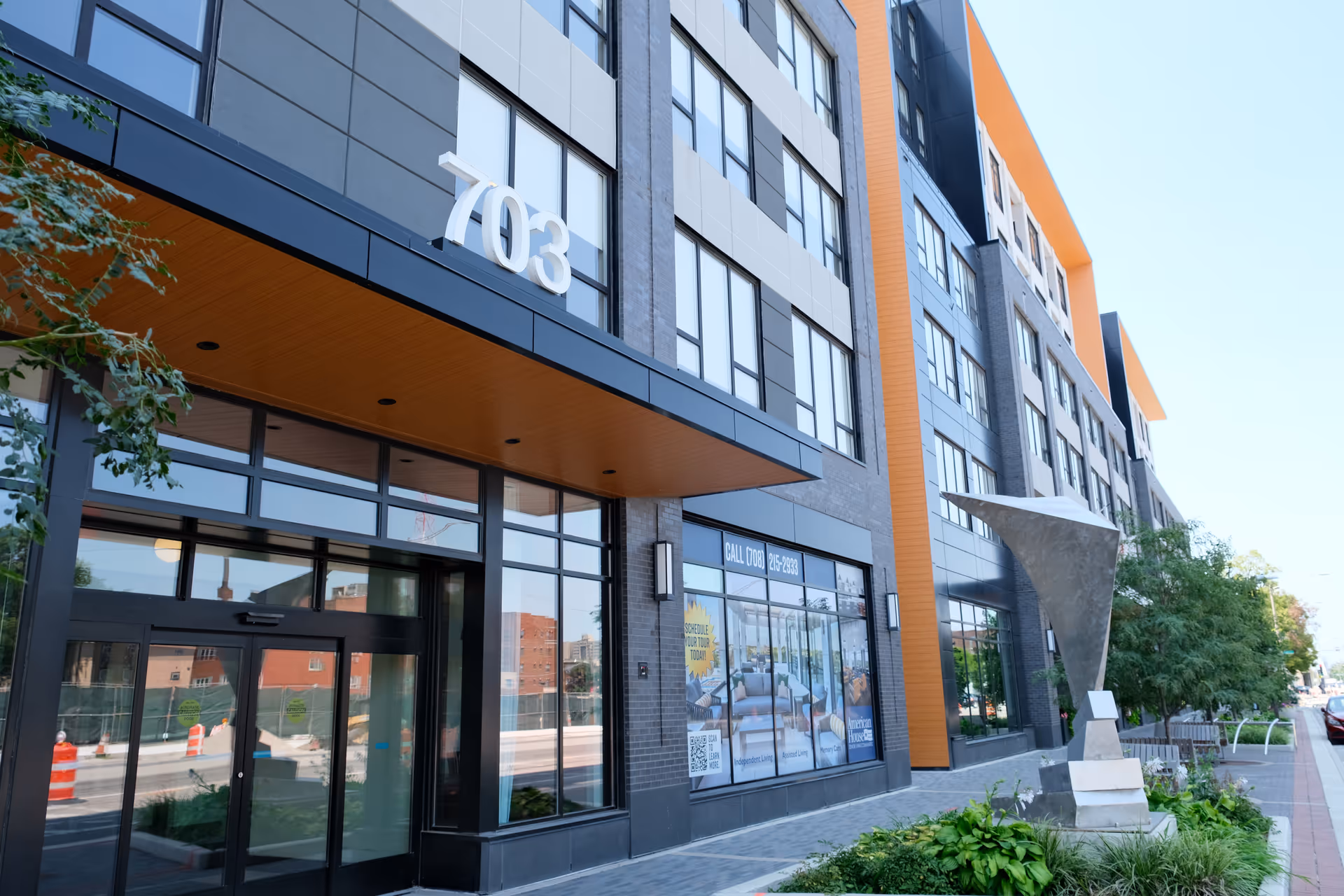Exterior view of a modern multi-story building with large windows and the number 703 displayed prominently above the entrance. There is a sidewalk with greenery and a metal sculpture near the entrance, and a clear blue sky in the background.
