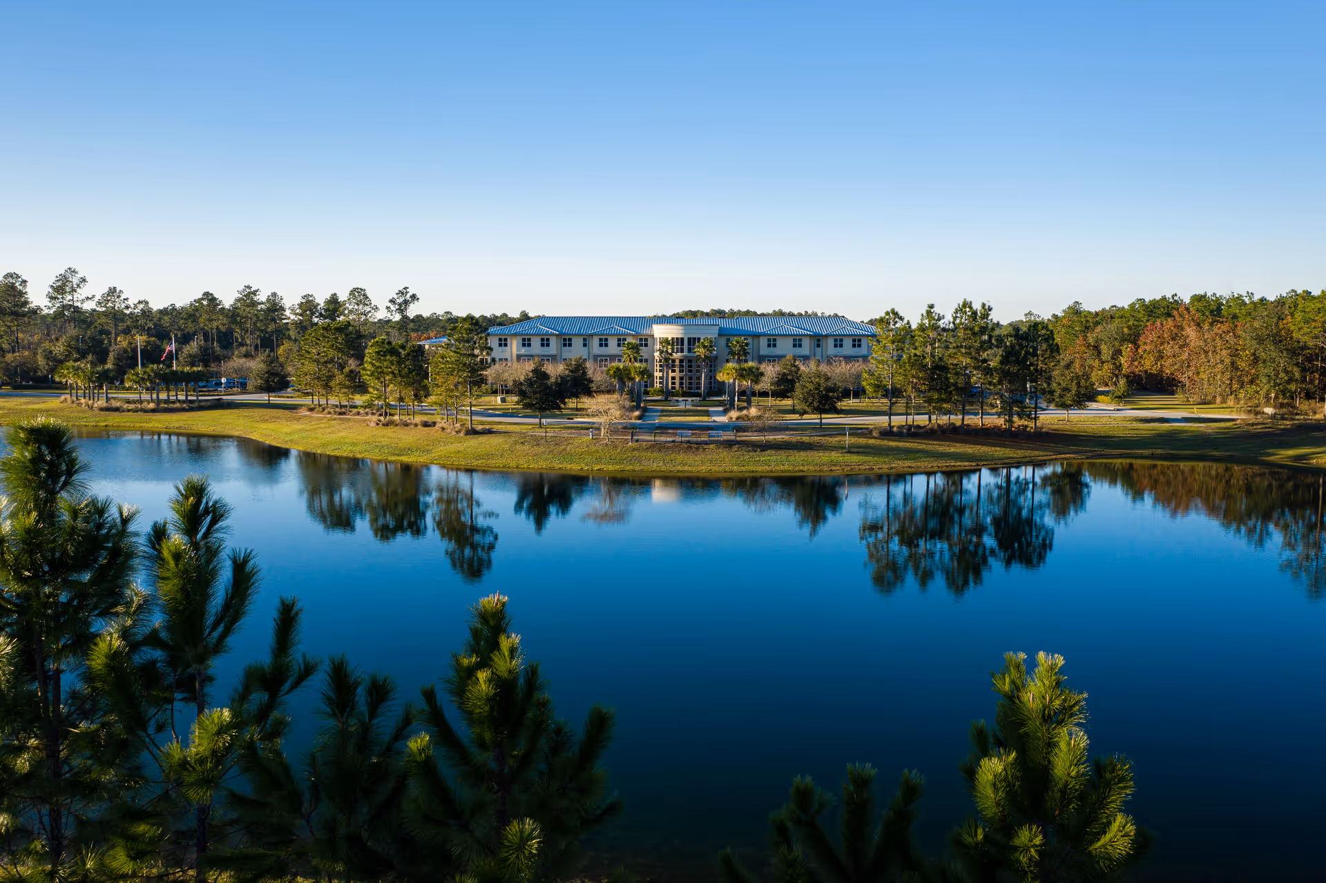A large building with a blue roof is situated behind a calm pond surrounded by trees and greenery under a clear blue sky.