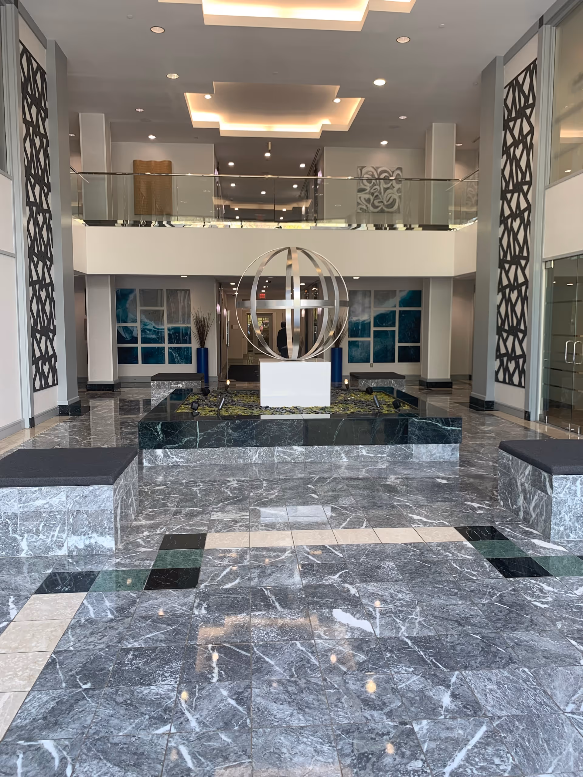 Modern, spacious lobby with marble floors, glass railings, and a spherical metal sculpture.