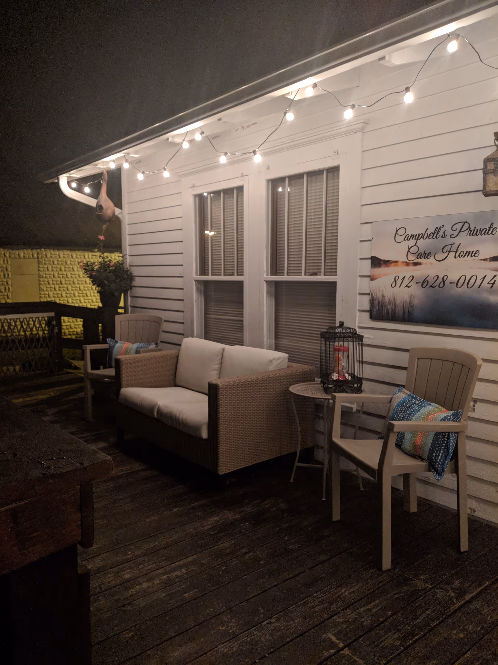 Outdoor wooden deck seating under string lights beside Campbell's Private Care Home sign.