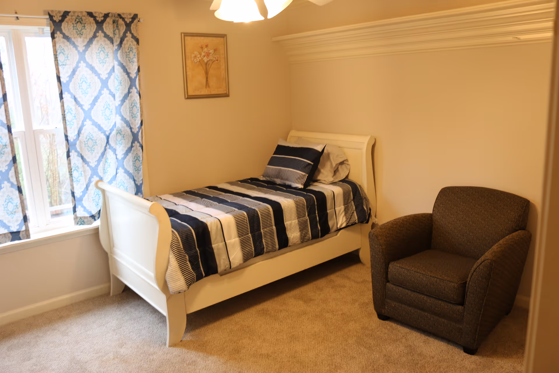 A small bedroom features a white single bed, blue curtains, and a brown armchair.