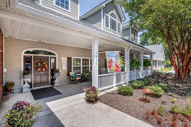 Front entrance of a senior living facility with a covered porch, decorated with a colorful welcome flag, potted plants, and a wreath on the door. The building has beige siding, white columns, and large windows, with a landscaped garden area to the right.