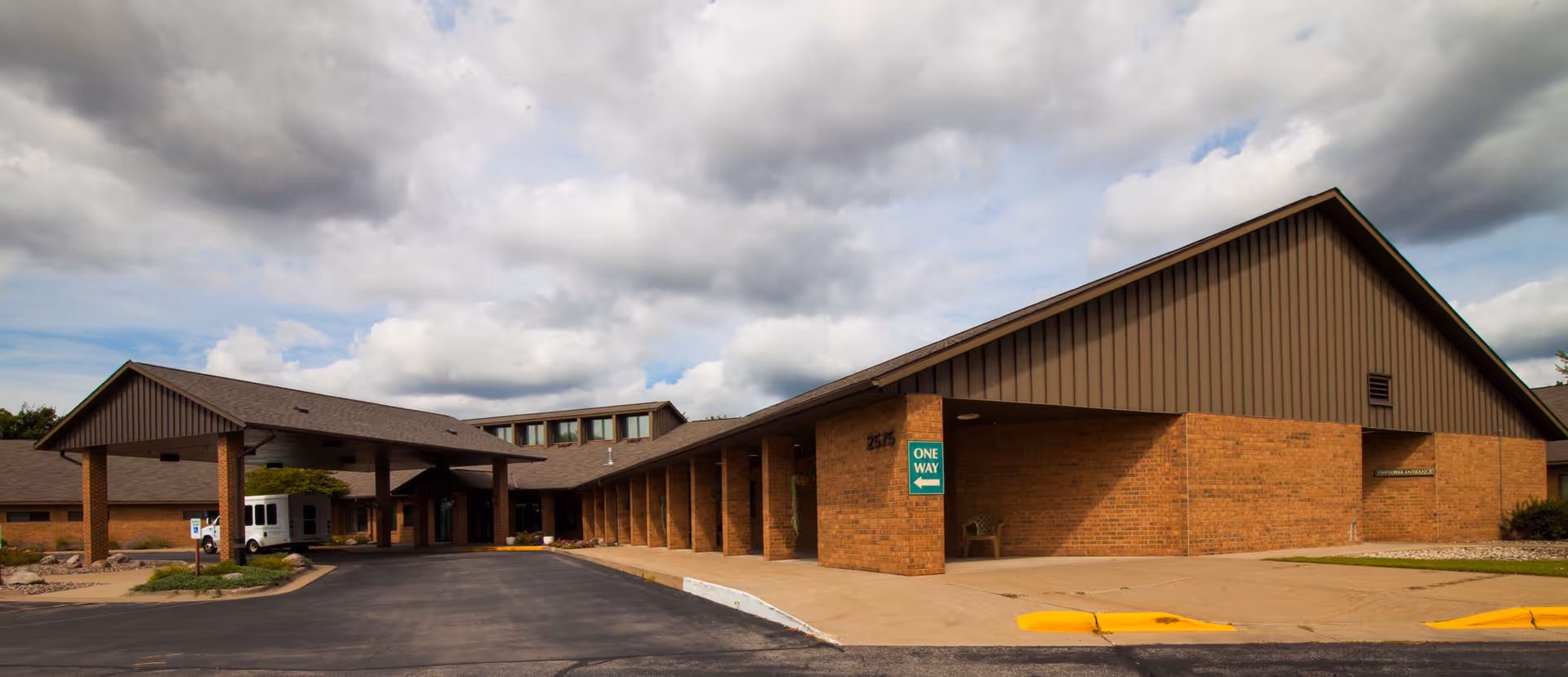 Exterior view of a single-story brick building with a covered entrance and a driveway. The building has a brown roof and siding, with a sign indicating 'One Way' and an 'Employee Entrance' sign visible. The sky is partly cloudy.