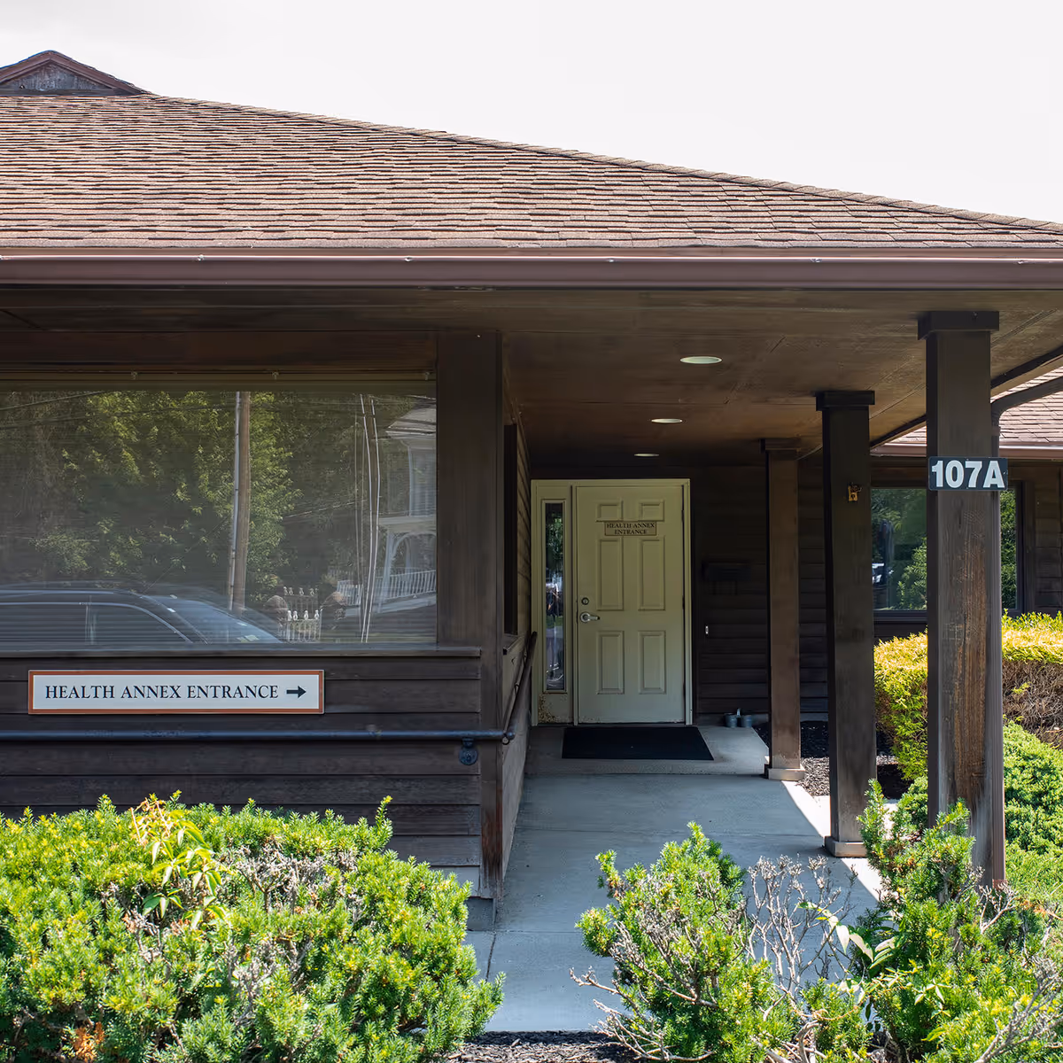 Front view of a wooden Health Annex entrance with a sign and surrounding shrubs.